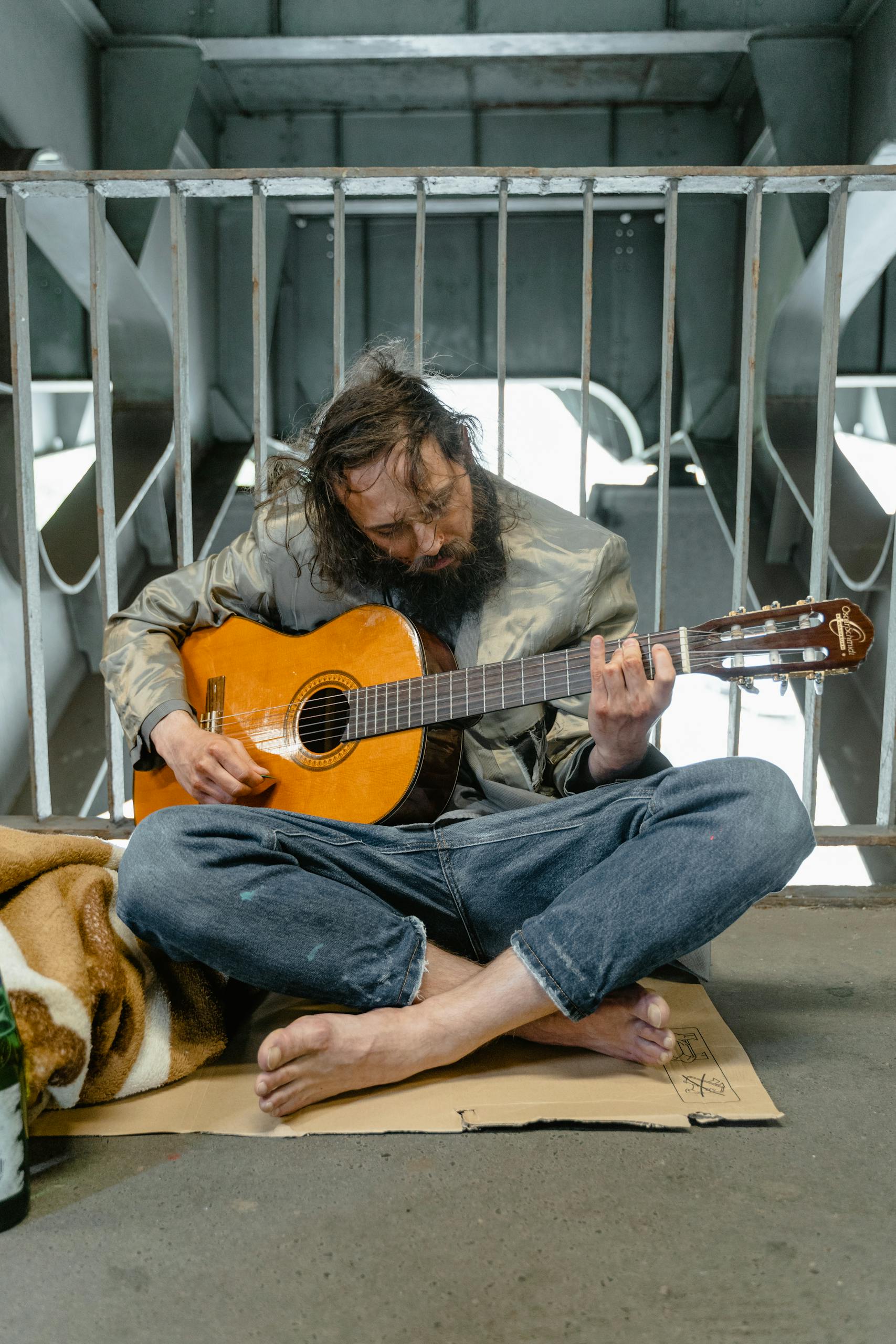 A man with a beard plays a guitar while sitting on cardboard in an urban environment.